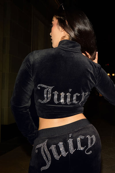 At night, a woman poses in a black Cropped Mockneck Jacket and matching pants, both with sparkling rhinestone Juicy logos on the back of the jacket and the seat of the pants.
