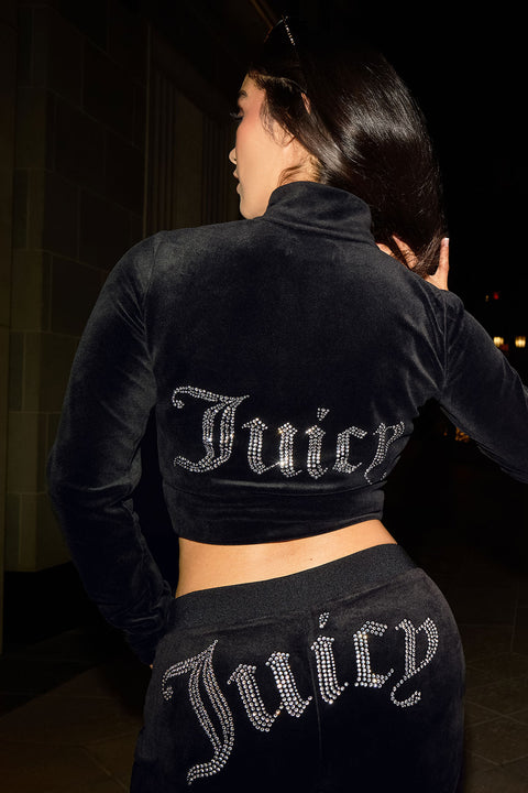 At night, a woman poses in a black Cropped Mockneck Jacket and matching pants, both with sparkling rhinestone Juicy logos on the back of the jacket and the seat of the pants.