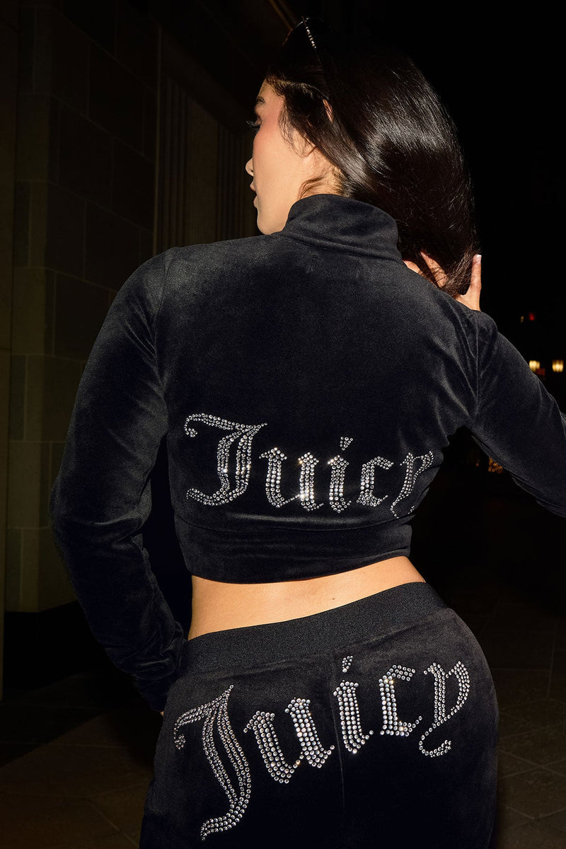 Wearing a Cropped Mockneck Jacket with a rhinestone Juicy logo on the back and matching pants, a person stands at night facing away from the camera.