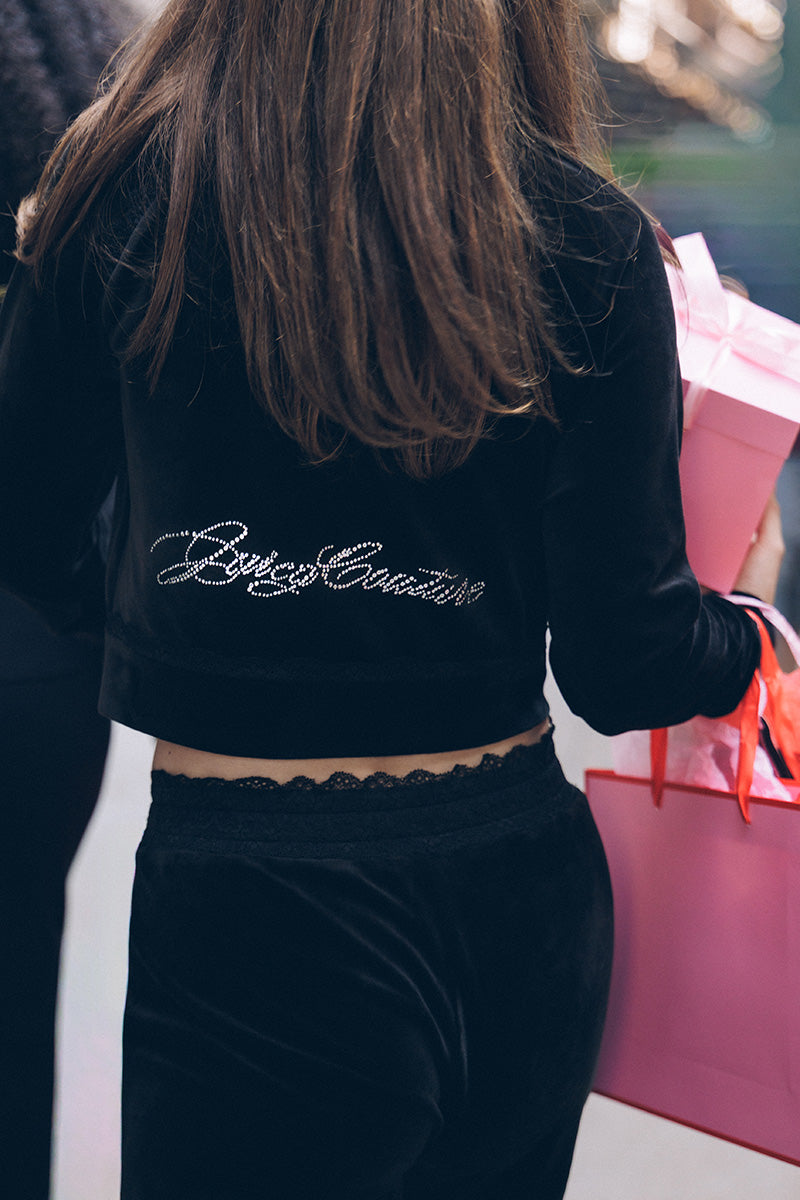 A person seen from behind wearing a Small Bling Velour Jacket, a black Y2K-inspired velour zip-up with script text spelled out in rhinestones on the back.