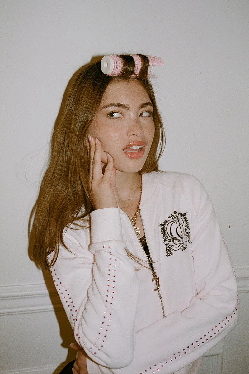 A person wearing a pale pink Scottie Crown Cotton Velour Jacket featuring a crest logo on the chest and rhinestone detailing along the sleeves.