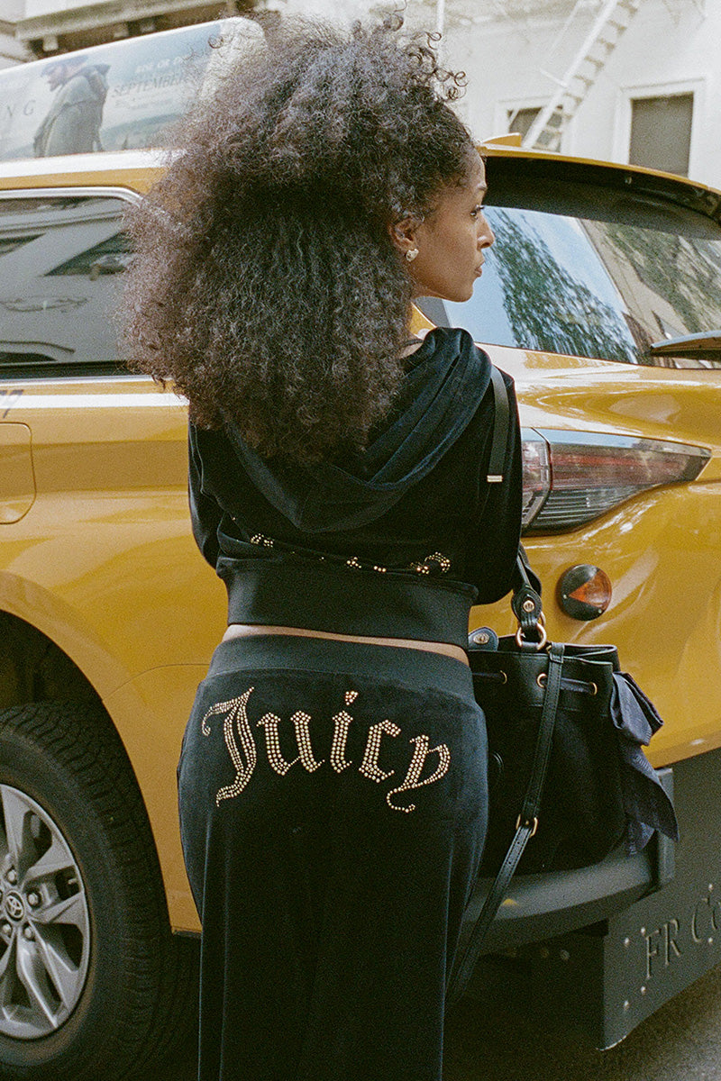 A person wearing black velour OG Big Bling Ribbon Track Pants with the word Juicy spelled out in rhinestones and ribbon details on the back, standing next to a yellow taxi.