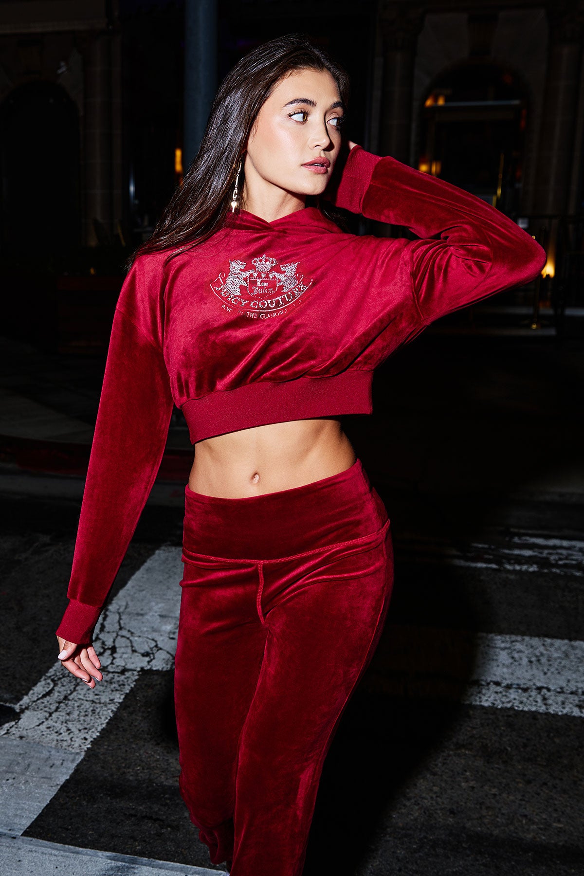 A woman walks on a city street at night, wearing a red velour matching set featuring the Cropped Scottie Hoodie and high-waisted pants, looking to the side with one hand touching her hair.