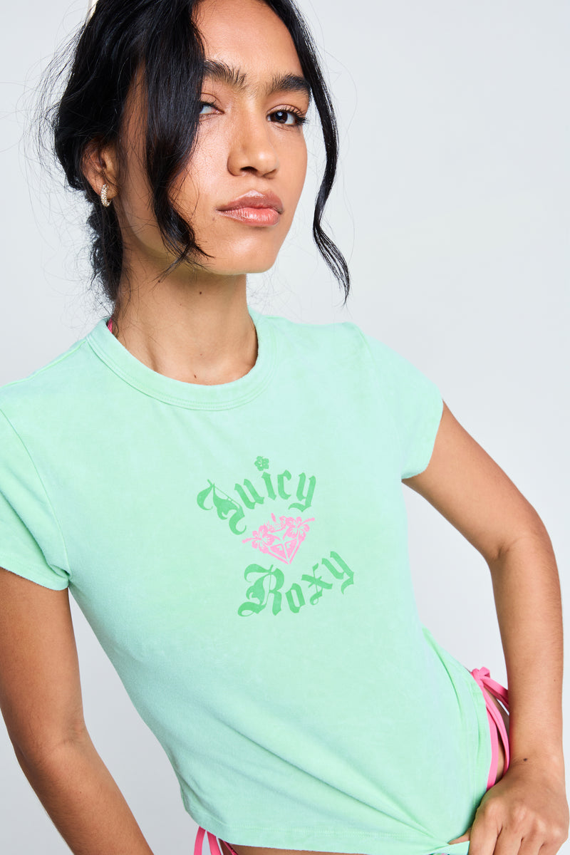 A woman with long dark hair wears a fitted short sleeve Roxy x Juicy Baby Tee in green with a pink heart, standing against a plain light background and looking confidently at the camera.