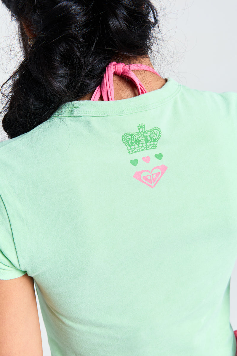 A person with long dark hair wears a fitted Roxy x Juicy Baby Tee in green, featuring a crown, three hearts, and a pink heart on the upper back, paired with visible pink halter straps around the neck.