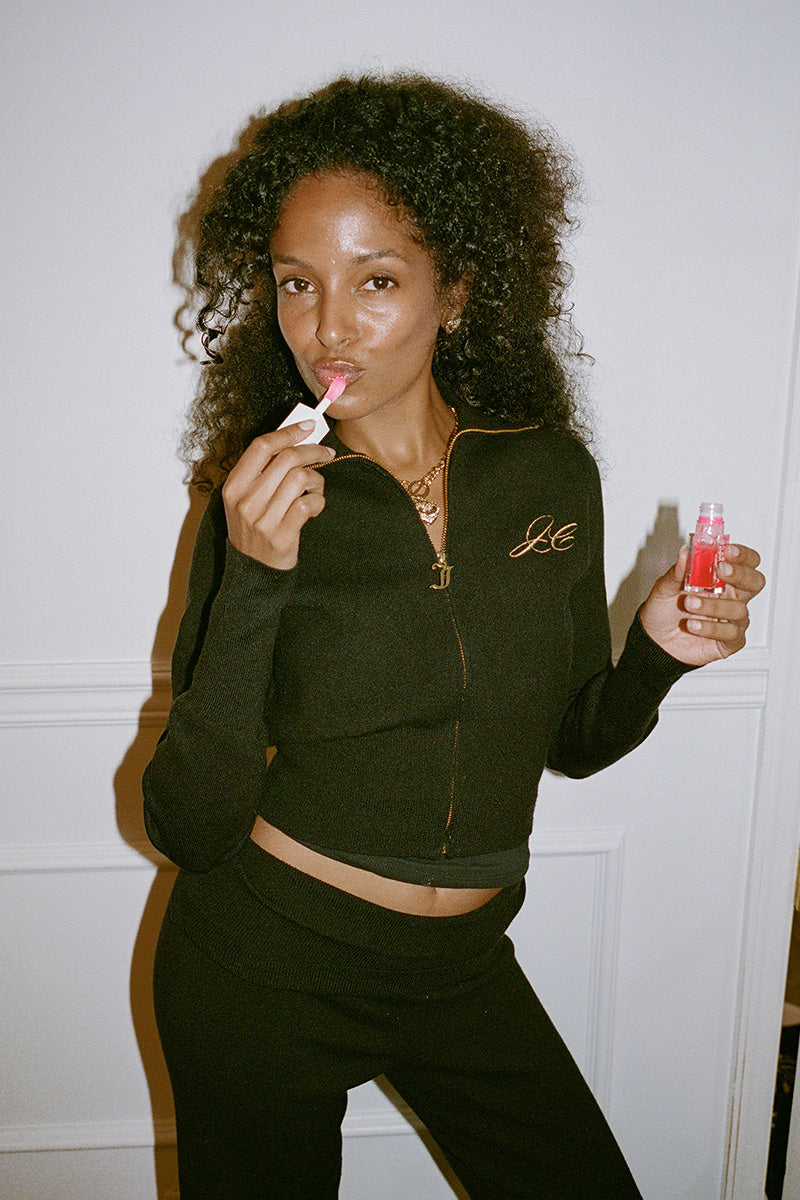 A woman wearing a black Embroidered Knit Cropped Jacket applies pink lip gloss.