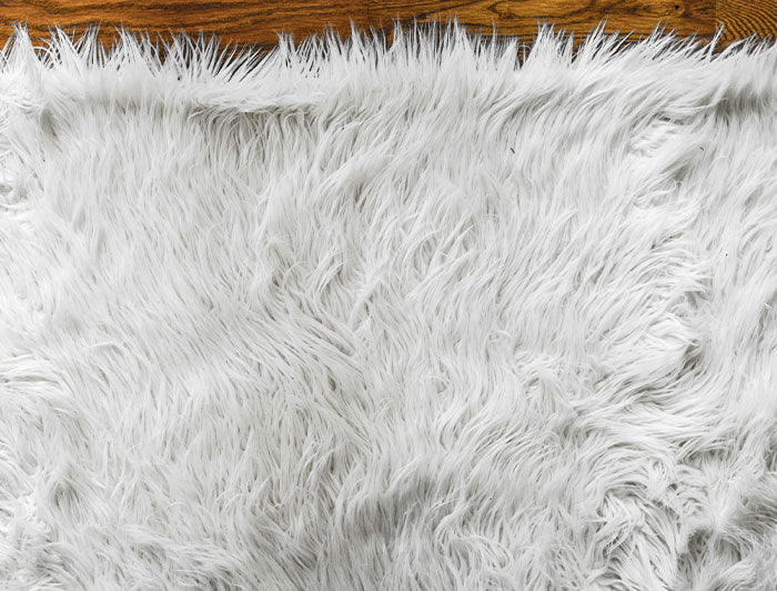A close-up of a white shaggy faux fur rug or blanket, showing its fluffy texture, with a small portion of a brown wooden floor visible at the top edge.