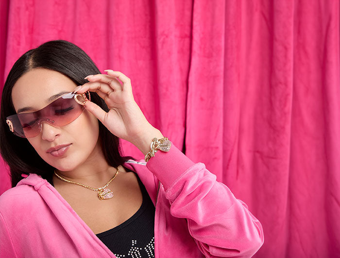 A woman wearing pink-tinted sunglasses with a gold frame, a pink hoodie, and gold jewelry, posing in front of a bright pink velvet curtain.