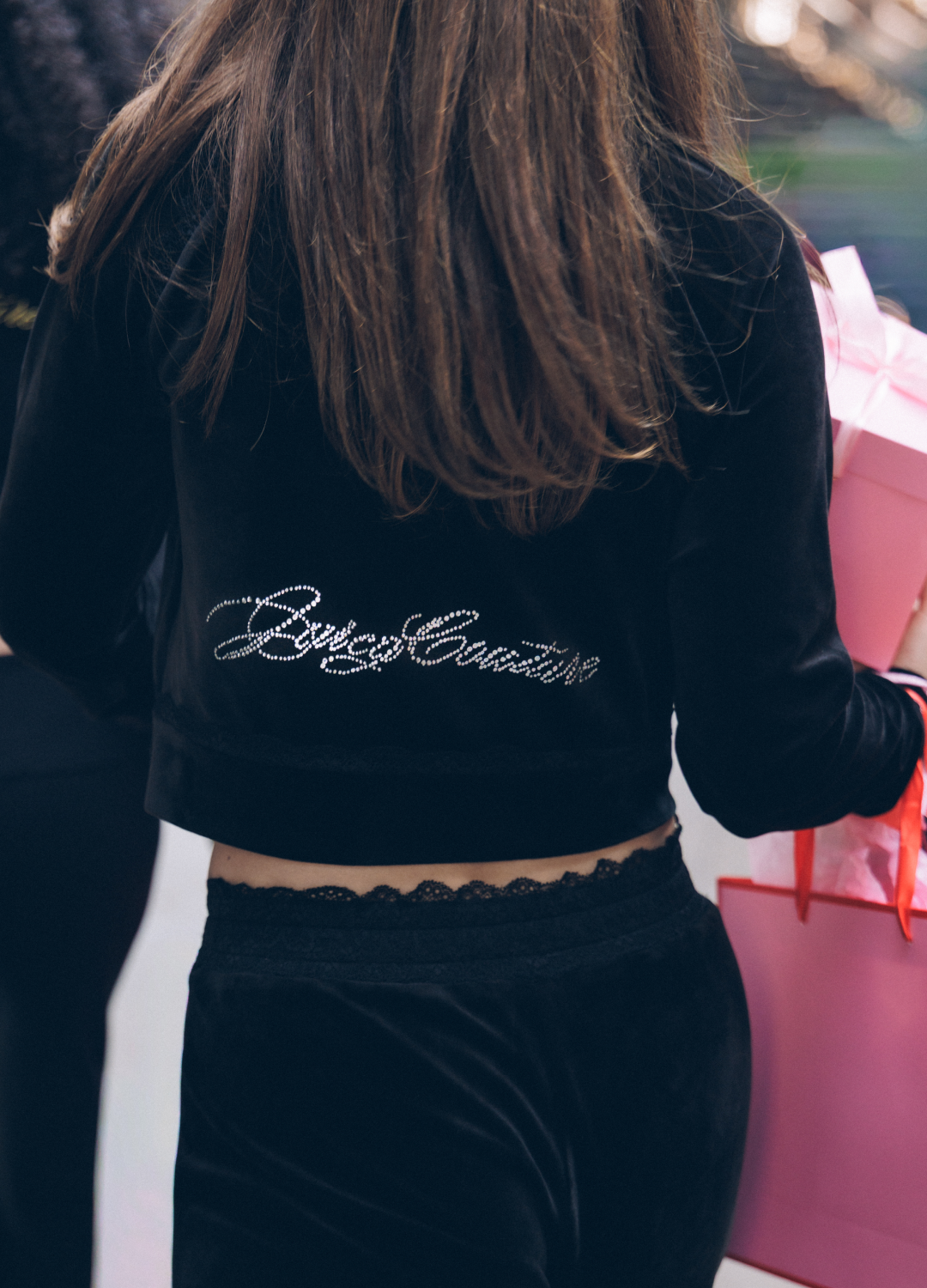 A person viewed from behind wearing a black velvet cropped sweatshirt with 'Juicy Couture' written across the back in silver script, paired with black lace-trimmed pants. The person is holding a pink shopping bag in an outdoor setting.