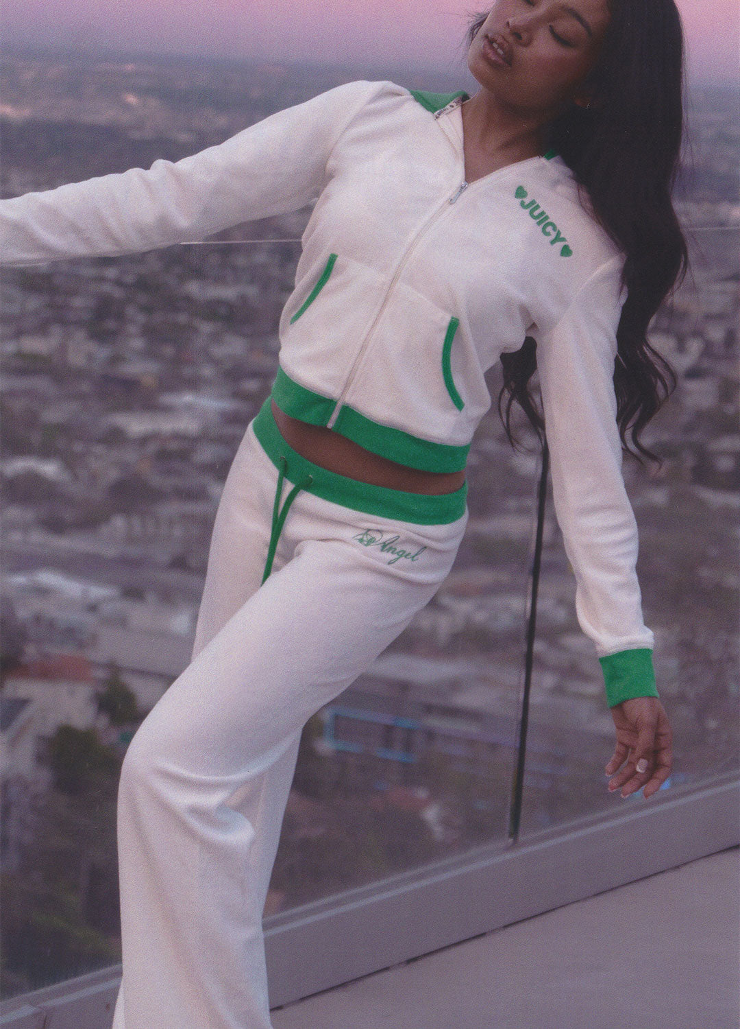 Person wearing a white and green tracksuit with 'Juicy' branding, standing on a balcony with a cityscape view.