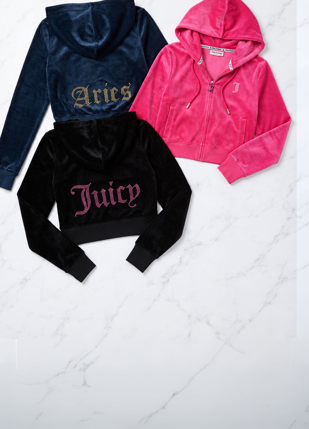 Three Juicy Couture cropped zip-up velour hoodies in navy blue, black, and pink, laid flat on a white marble background. Each hoodie features the brand's signature Gothic-style embroidered logo on the back.