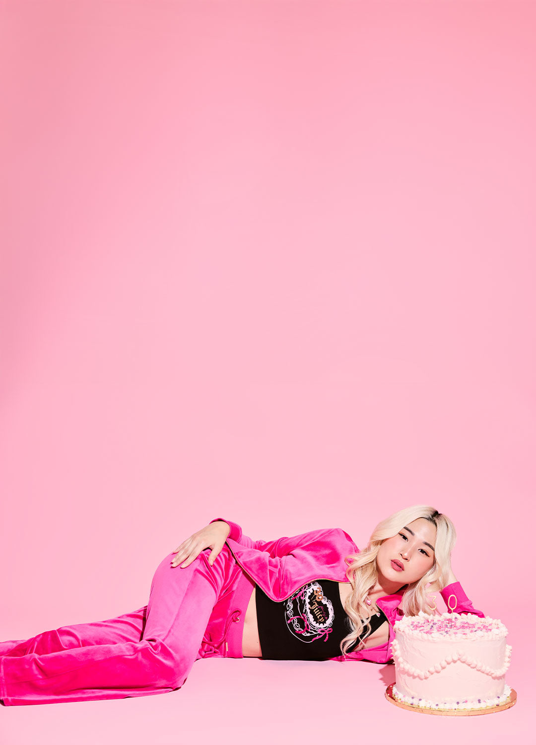 Person in pink tracksuit and black top with a cake on a pink background