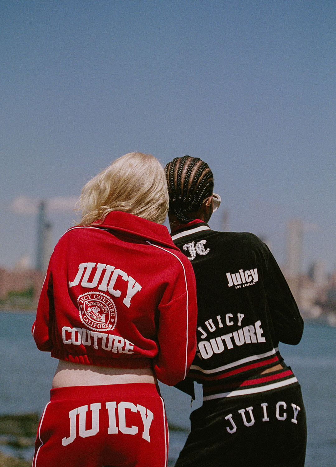 Two models from behind wearing Juicy Couture tracksuits, one in red and one in black, standing outdoors near a waterfront with a city skyline in the background.