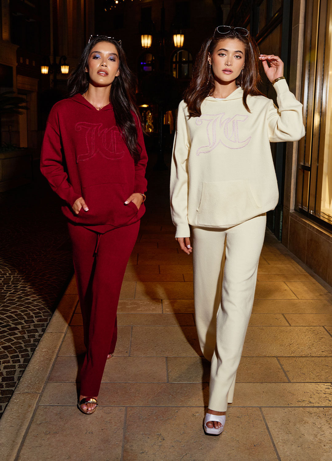 Two women in red and beige Juicy Couture tracksuits standing in a dimly lit street with decorative elements.