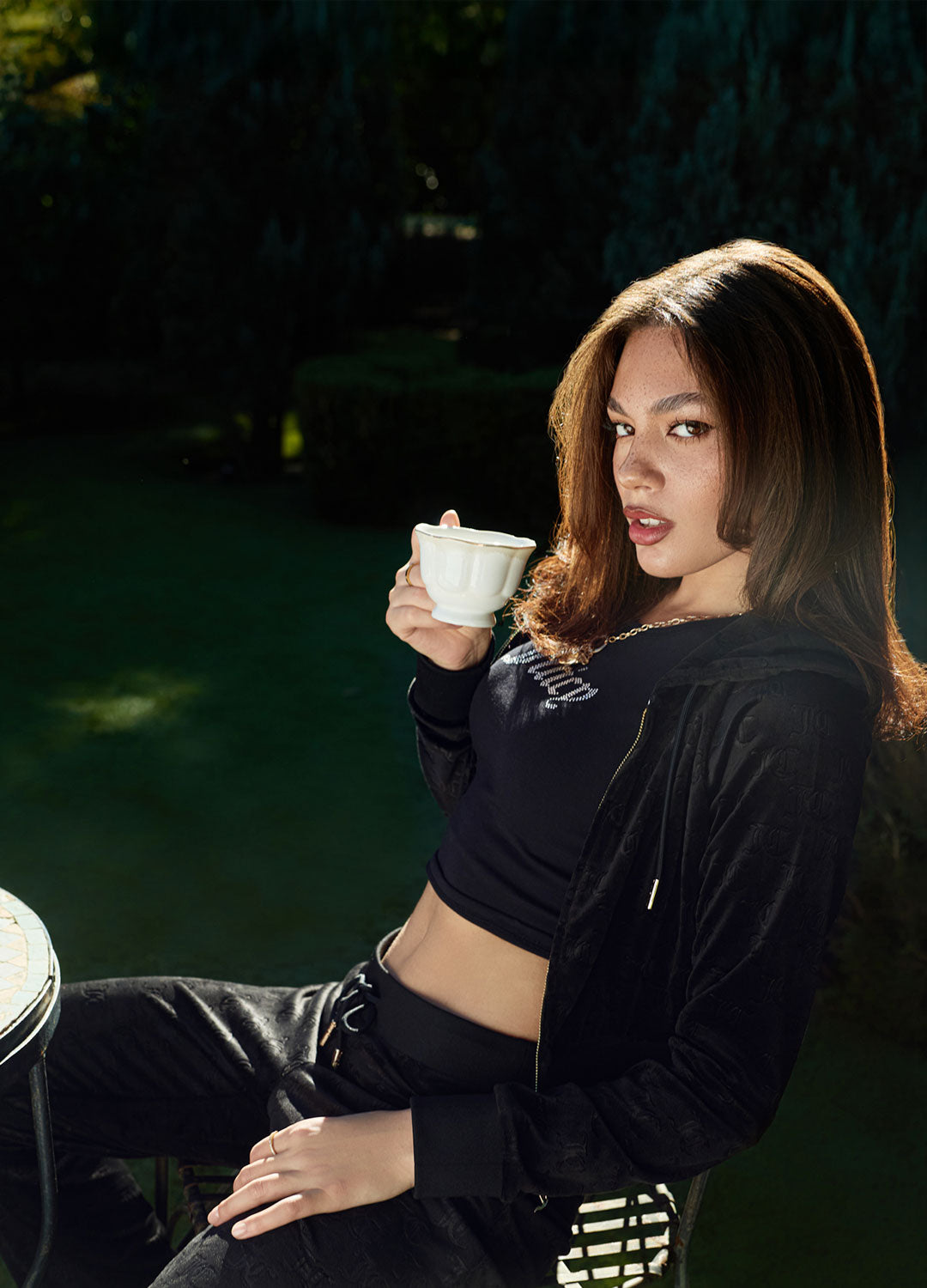 Woman in black tracksuit sitting outdoors by a table with a teapot and cups, holding a cup.