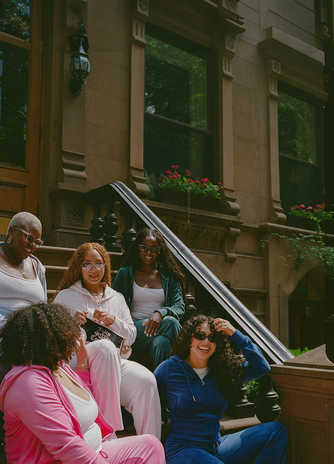 Five women in bright pink, light pink, blue, light blue, and green tracksuits sitting together on the steps of a brownstone building in the city.