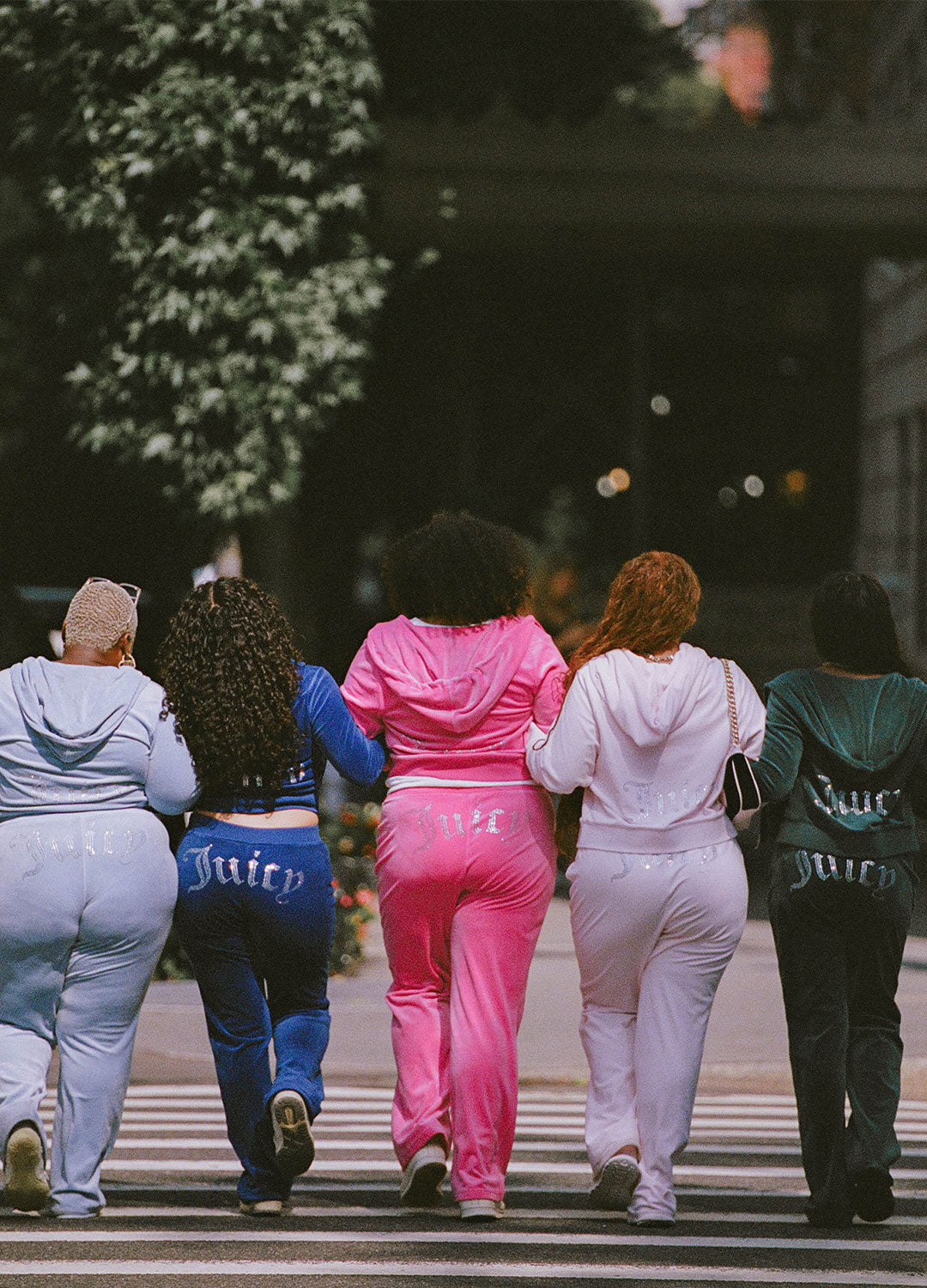 Five women walking hand-in-hand across a city street crosswalk, viewed from behind. Each is wearing a matching Juicy Couture tracksuit in a different color: light blue, dark blue, pink, light pink, and dark green.