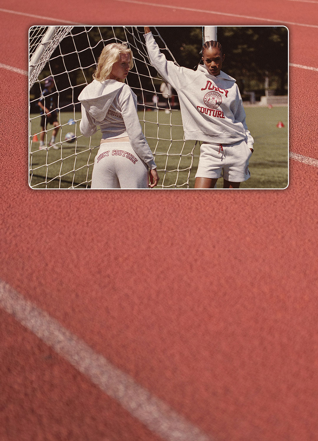 A lifestyle photo of two women on a track field wearing a matching light gray athletic tracksuit with red lettering, set against a red running track background with a soccer goal net in the foreground.