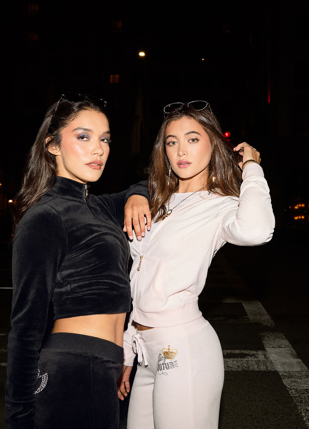 Two women in a light pink Juicy Couture tracksuit and a black Juicy Couture tracksuit posing in a street at night.