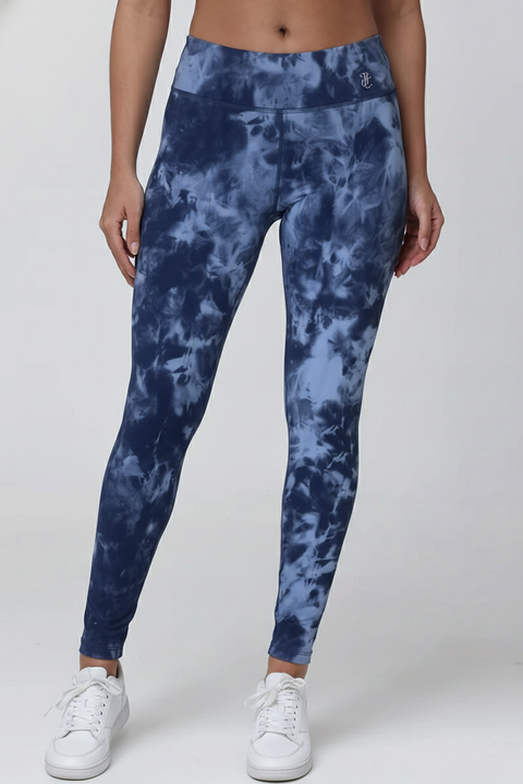 A person wears Lorraine Printed Legging—REJUICED, featuring a high-waisted blue tie-dye design, paired with white sneakers. Only the lower torso and legs are shown against a plain light background.