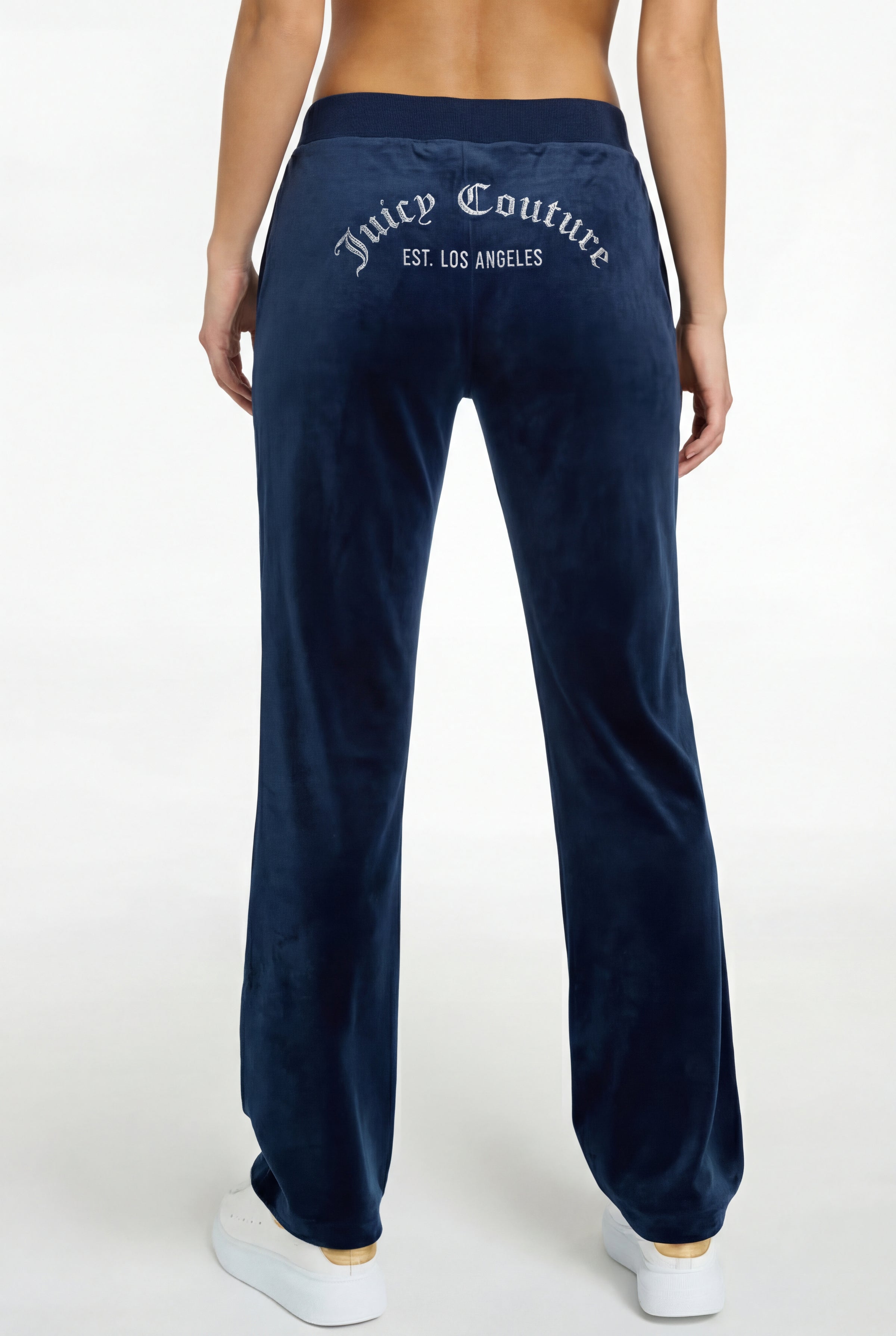 Someone is seen from the back wearing Arched Diamante Classic Velour Trackpant—REJUICED in navy blue, featuring “Juicy Couture EST. LOS ANGELES” in silver across the waistband, paired with white sneakers.