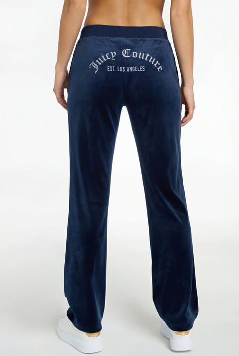 Someone is seen from the back wearing Arched Diamante Classic Velour Trackpant—REJUICED in navy blue, featuring “Juicy Couture EST. LOS ANGELES” in silver across the waistband, paired with white sneakers.
