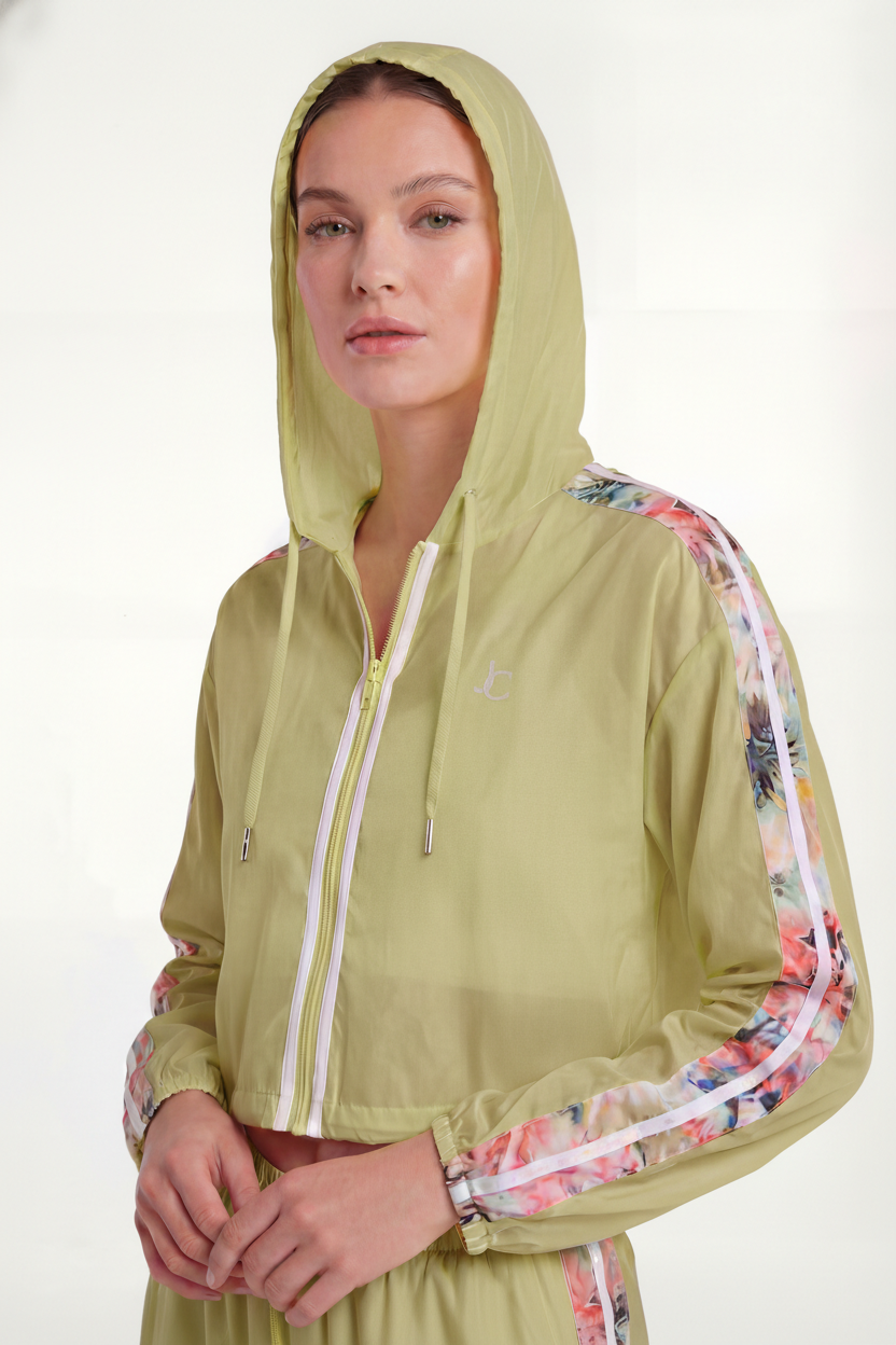 A woman wears the REJUICED Long Sleeve Cropped Hooded Zip Front jacket in light green with floral sleeve stripes and the hood up, standing against a plain light background.