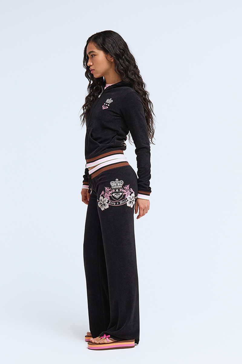 Woman in profile wears Roxy x Juicy OG Straight Pants, a Y2K style black velour tracksuit with ultra low rise, embroidered crest and floral designs, paired with pink flip-flops; plain white background.
