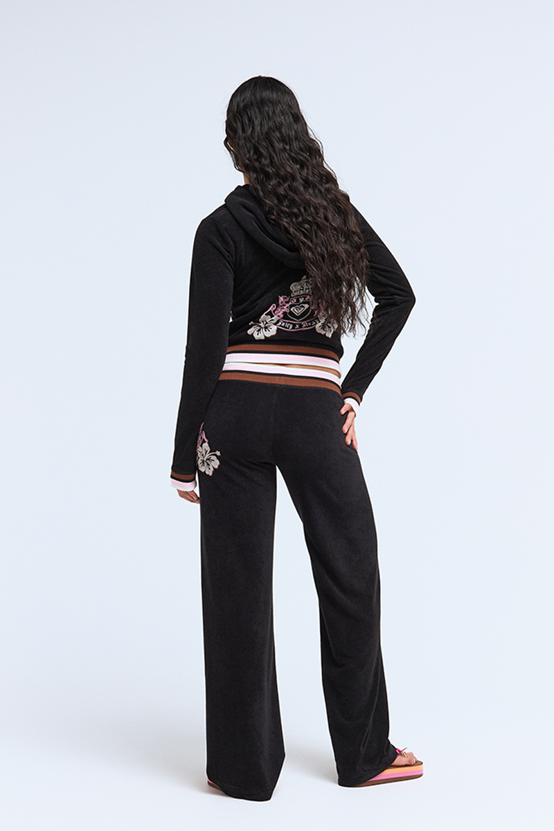 A person with long, wavy hair faces away in Roxy x Juicy OG Straight Pants featuring black velour and floral back/hip embroidery, paired with pink flip-flops, channeling Y2K style against a plain light background.
