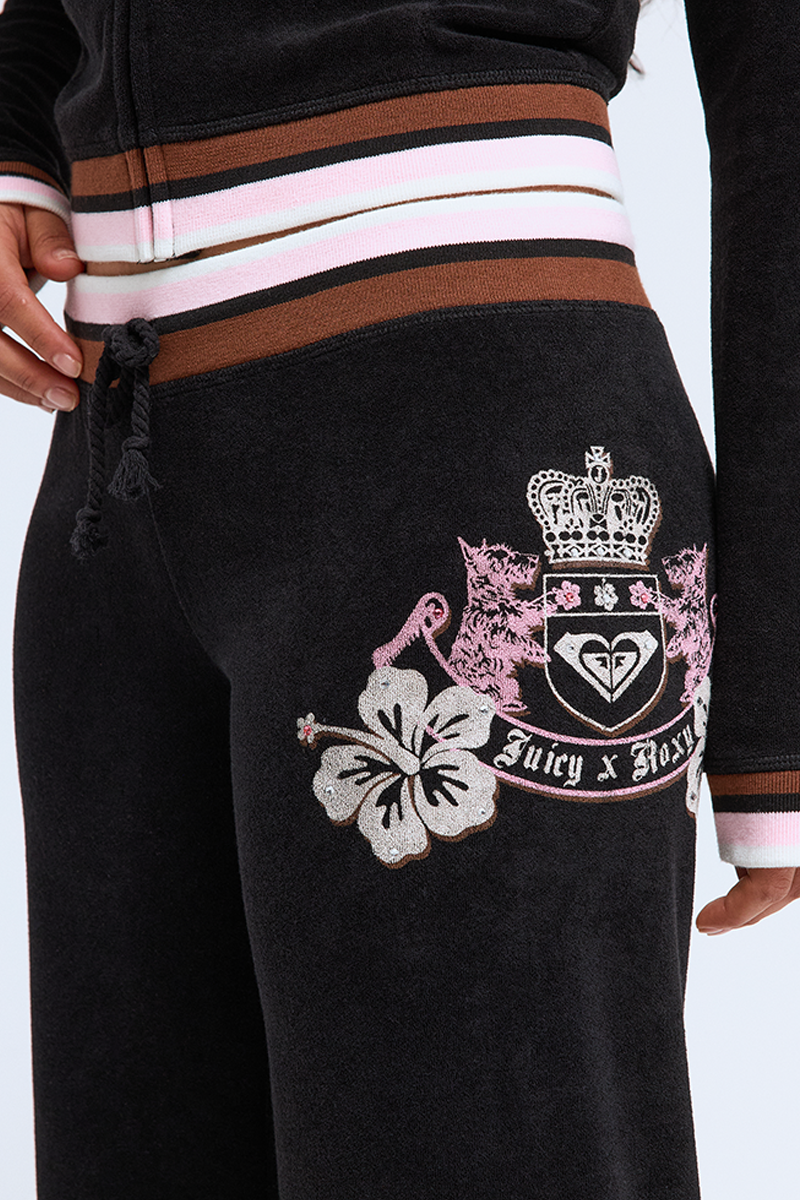 Close-up of Roxy x Juicy OG Straight Pants in black velour with a pink, white, and brown waistband and cuffs. Features a crest, pink floral details, and "Juicy x Roxy" text on the upper thigh for true Y2K style.