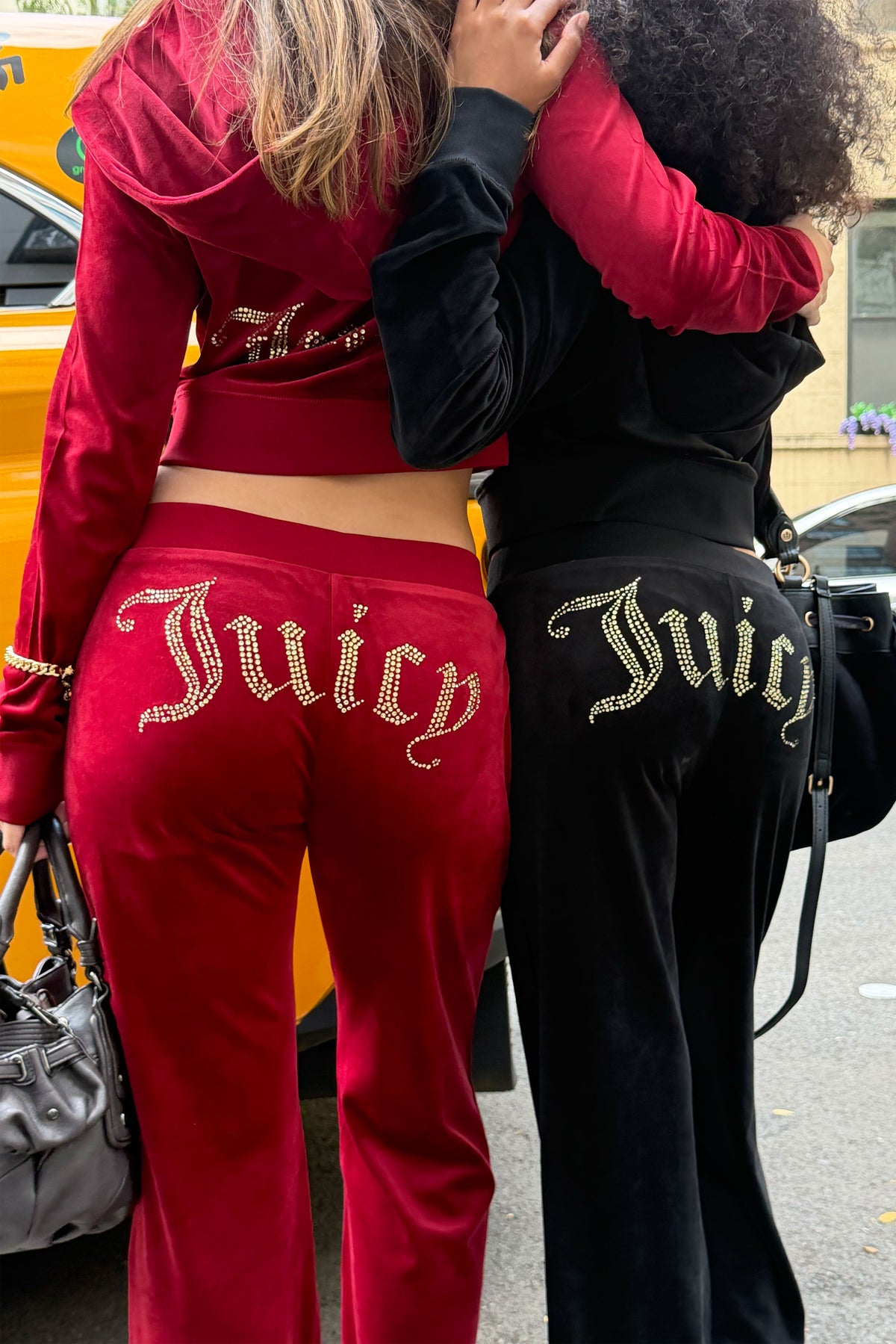 Two women standing outdoors in matching velvet tracksuits, shown from the back. One wears a red set with large gold rhinestone 'Juicy' lettering on the pants, and the other wears a black set with matching lettering.