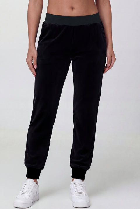 Someone wearing Black Velour Cinched Jogger Pants—REJUICED, featuring a dark waistband and cuffs, paired with white sneakers, stands against a plain light background.