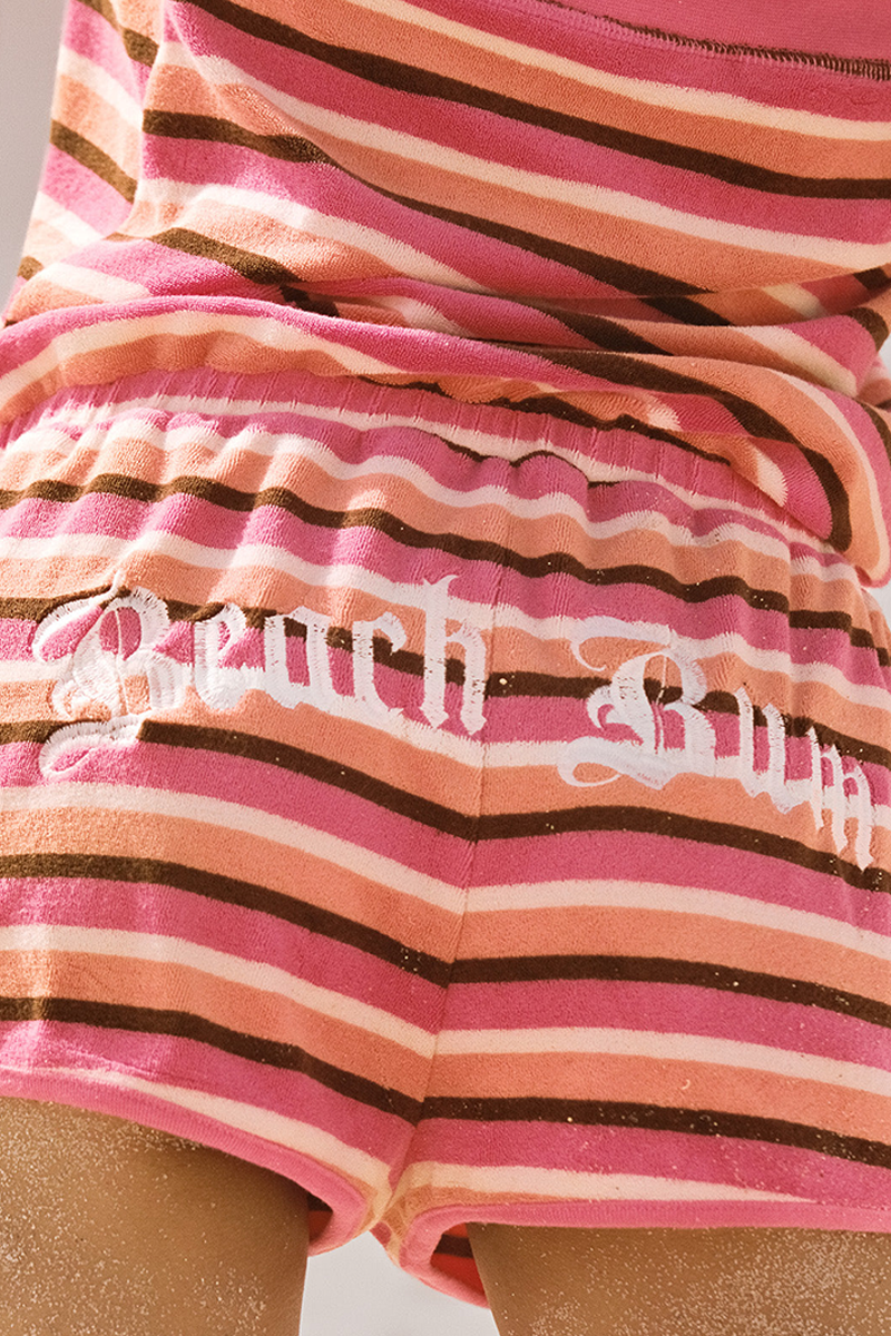 Someone wears the Roxy x Juicy Stripe Romper, featuring pink stripes and "Beach Bum" in white script across the back. Its soft, terry-towelling-like material has visible sand marking a beach-ready vibe.