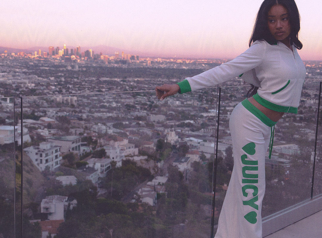 A woman modeling a white Juicy Couture tracksuit with green accents, featuring a cropped long-sleeve top and matching pants with 'JUICY' printed vertically, set against a city skyline at sunset.